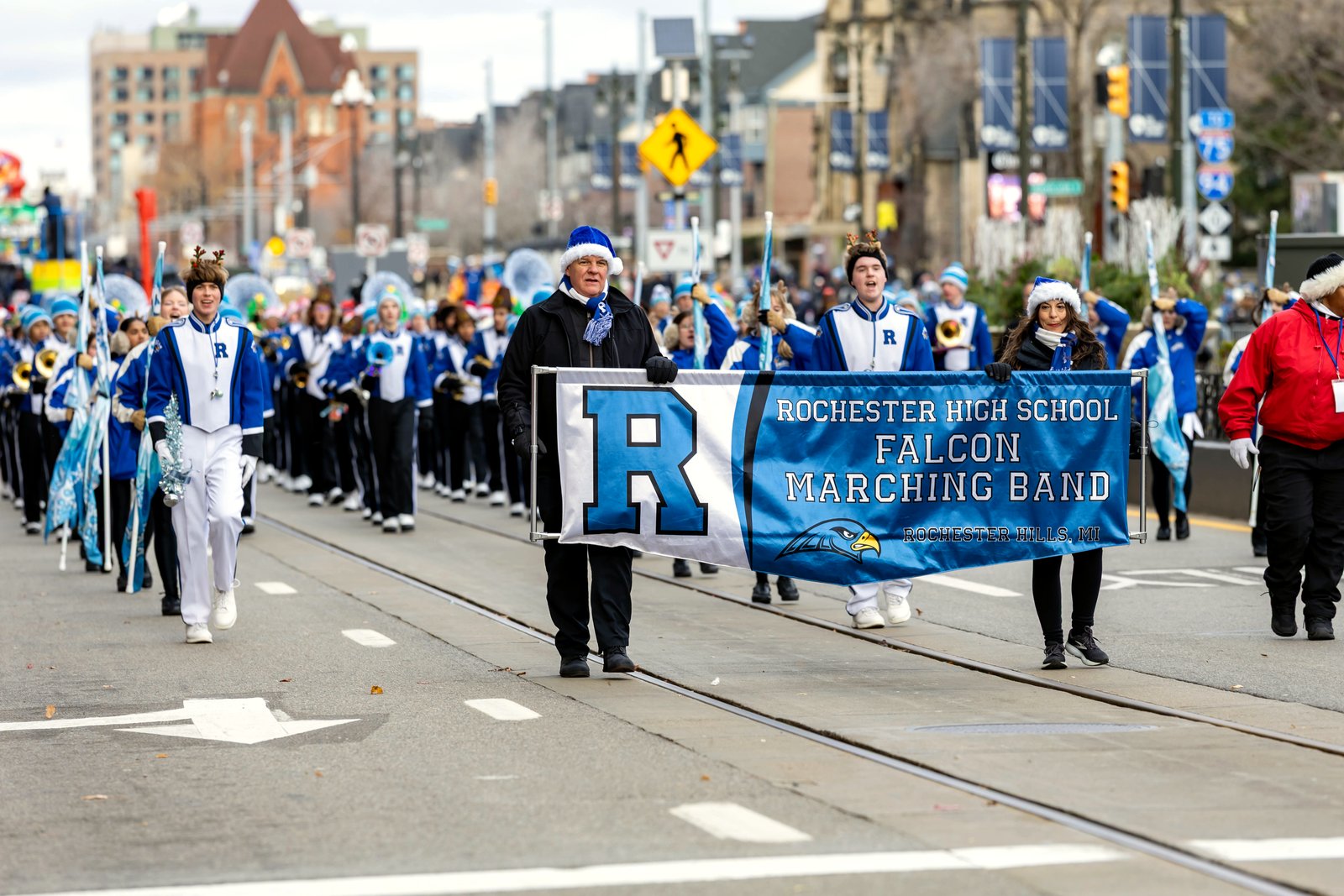 A high school marching band in blue and white uniforms parades down a city street behind a banner reading Rochester High School Falcon Marching Band.
