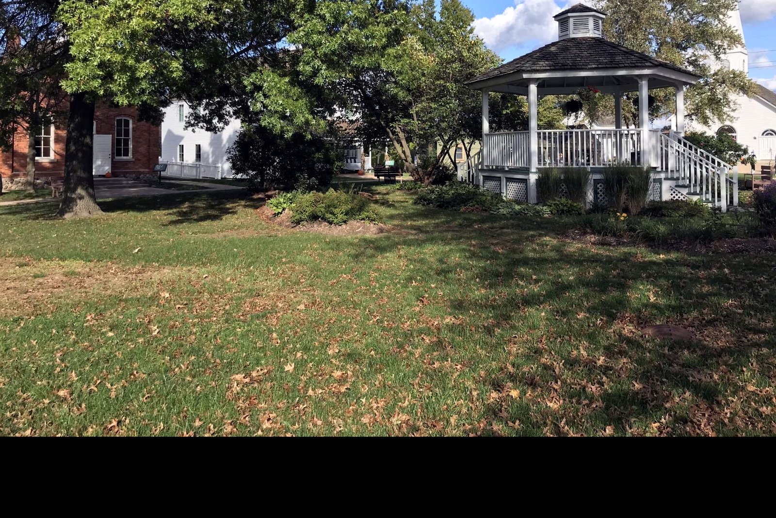 A white gazebo on a grassy village green framed by a large shade tree, with a red brick building and a white church steeple in the background.
