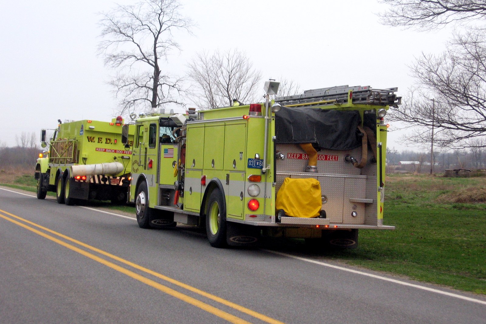 Two yellow-green fire trucks parked along the shoulder of a two-lane rural road beside a field and bare trees.