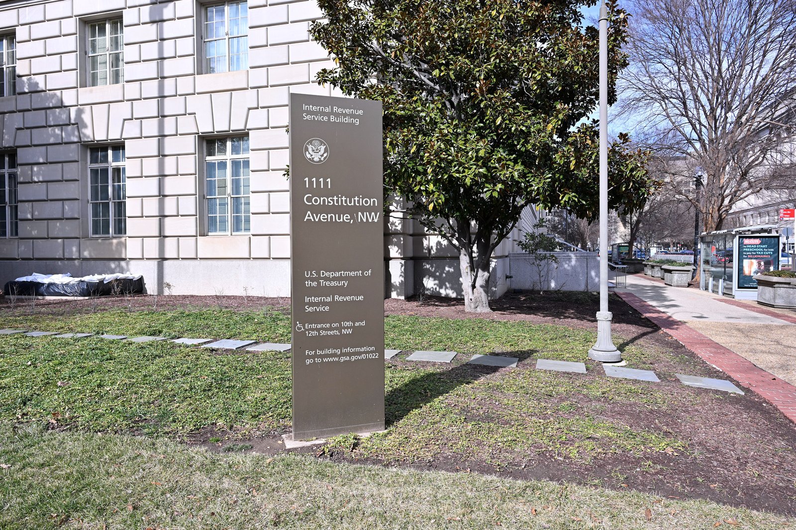 A brown sign reading Internal Revenue Service Building, 1111 Constitution Avenue NW, on a lawn beside a stone federal building and a magnolia tree.