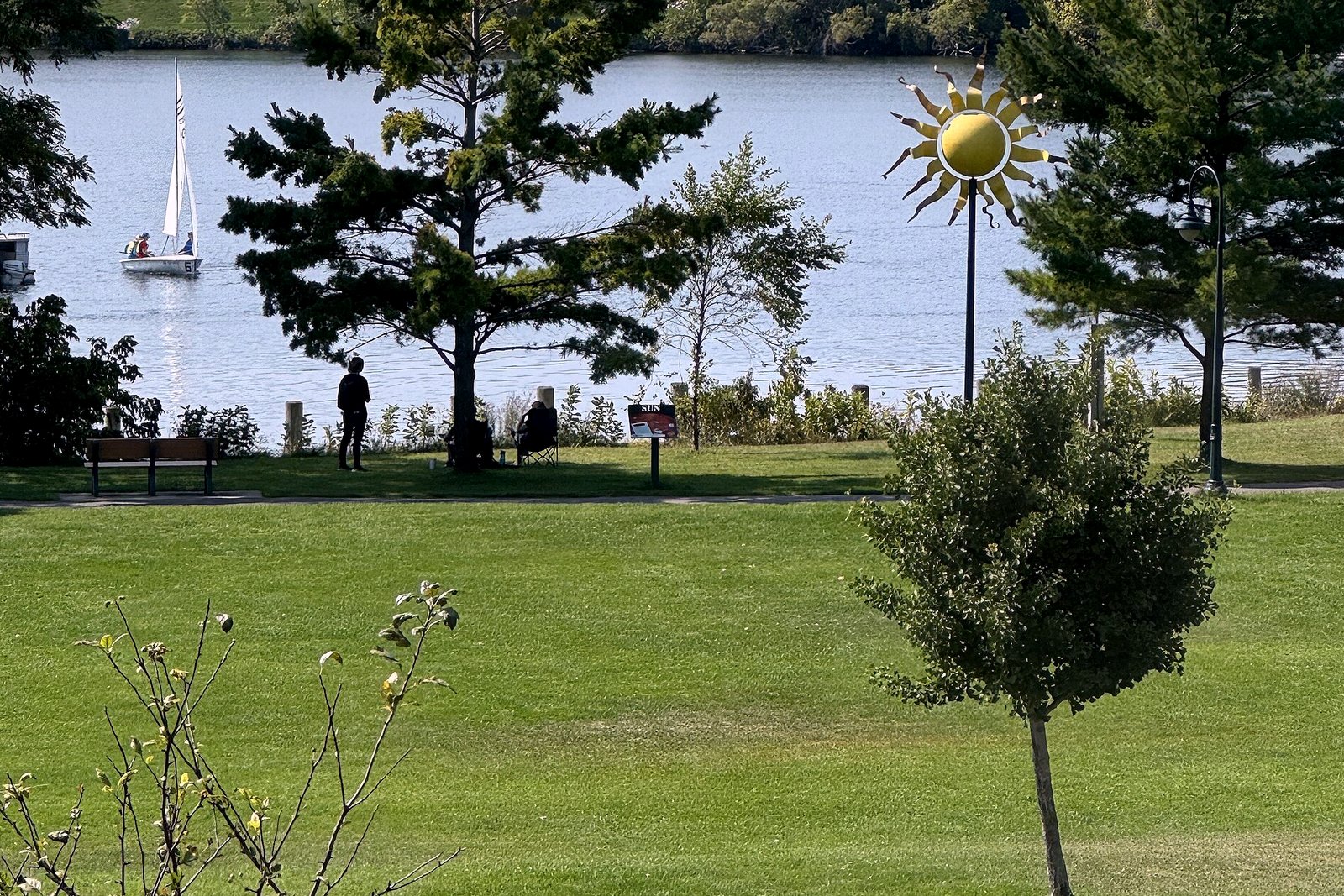 A grassy lakeside park with young trees and a sun-shaped sculpture on a pole, seen from above, with a sailboat on the lake and a wooded shoreline behind it.