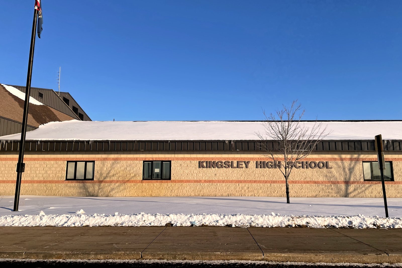 A tan brick school building with a sign reading Kingsley High School, a bare tree and a flagpole in front, under a deep blue winter sky.