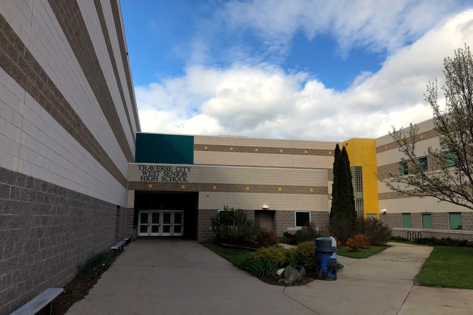 The entrance of a large tan-brick high school building, with glass doors, shrubs and a walkway under a partly cloudy sky.