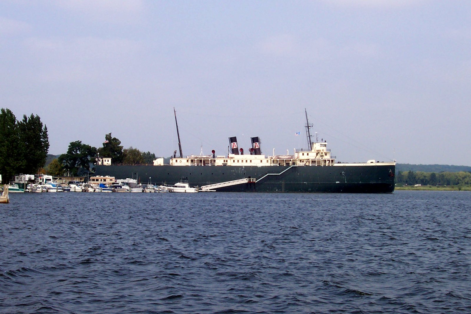 A long black-hulled retired car ferry with a white superstructure, docked on the far shore across open water.