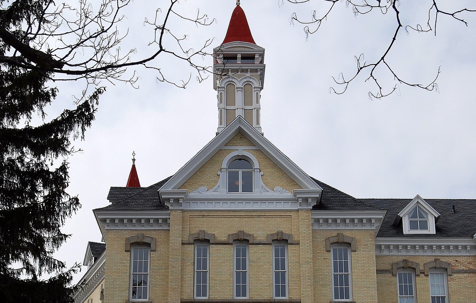 The cream-colored brick upper floors and central gable of a Victorian building, topped by a tall white cupola with a red spire, framed by bare tree branches and an evergreen against an overcast sky.