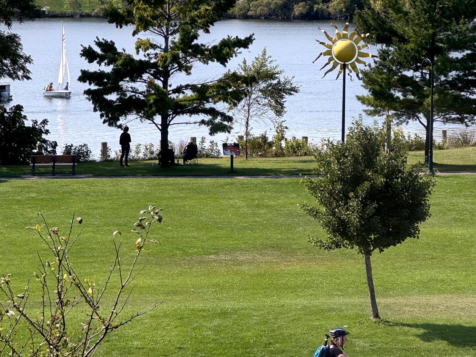 A grassy lakeside park with young trees, a sun-shaped sculpture and a sailboat on the water beyond, seen from above on a sunny day.