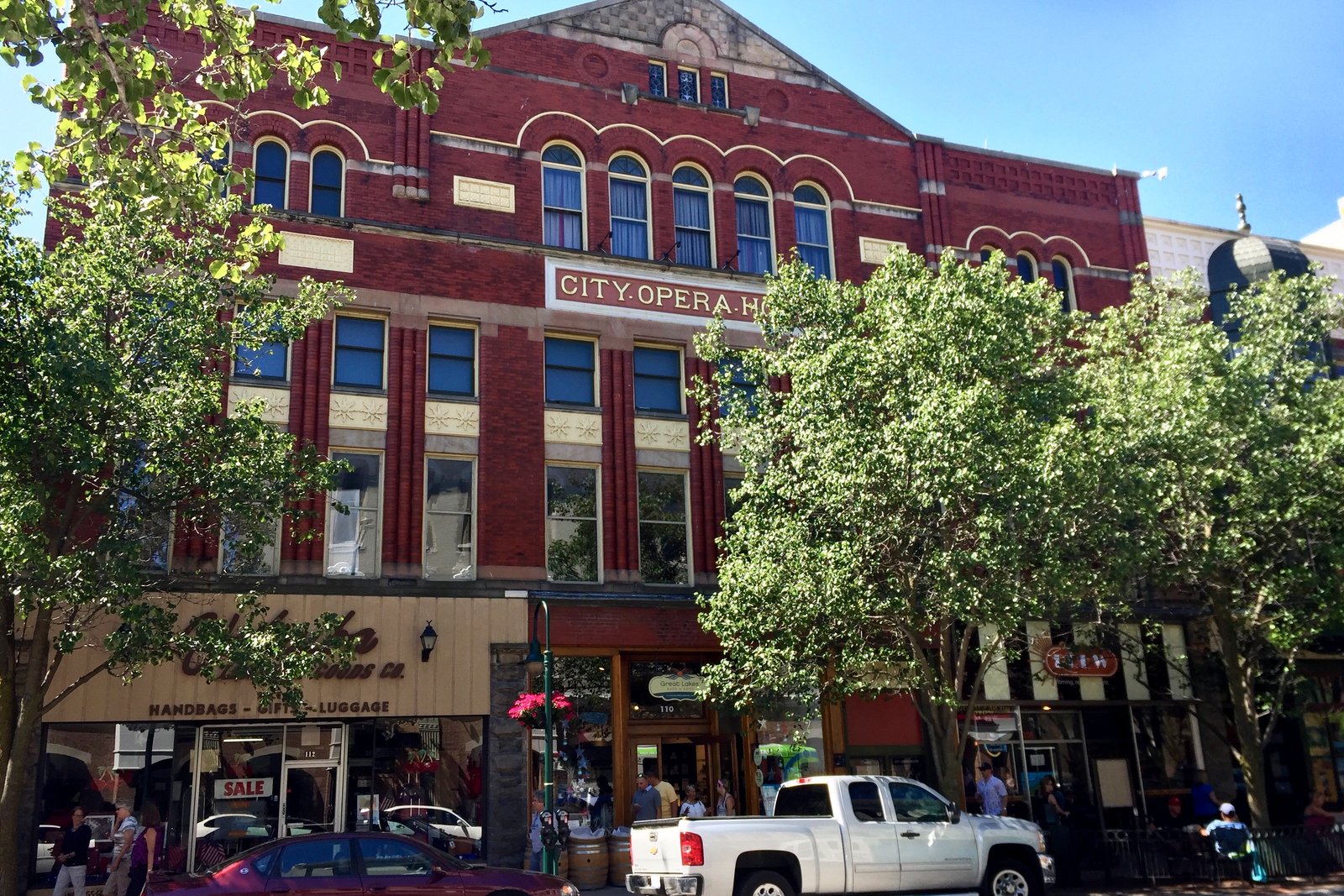 The red brick City Opera House on Front Street in Traverse City, with arched upper windows and its name across the facade, shaded by leafy street trees.