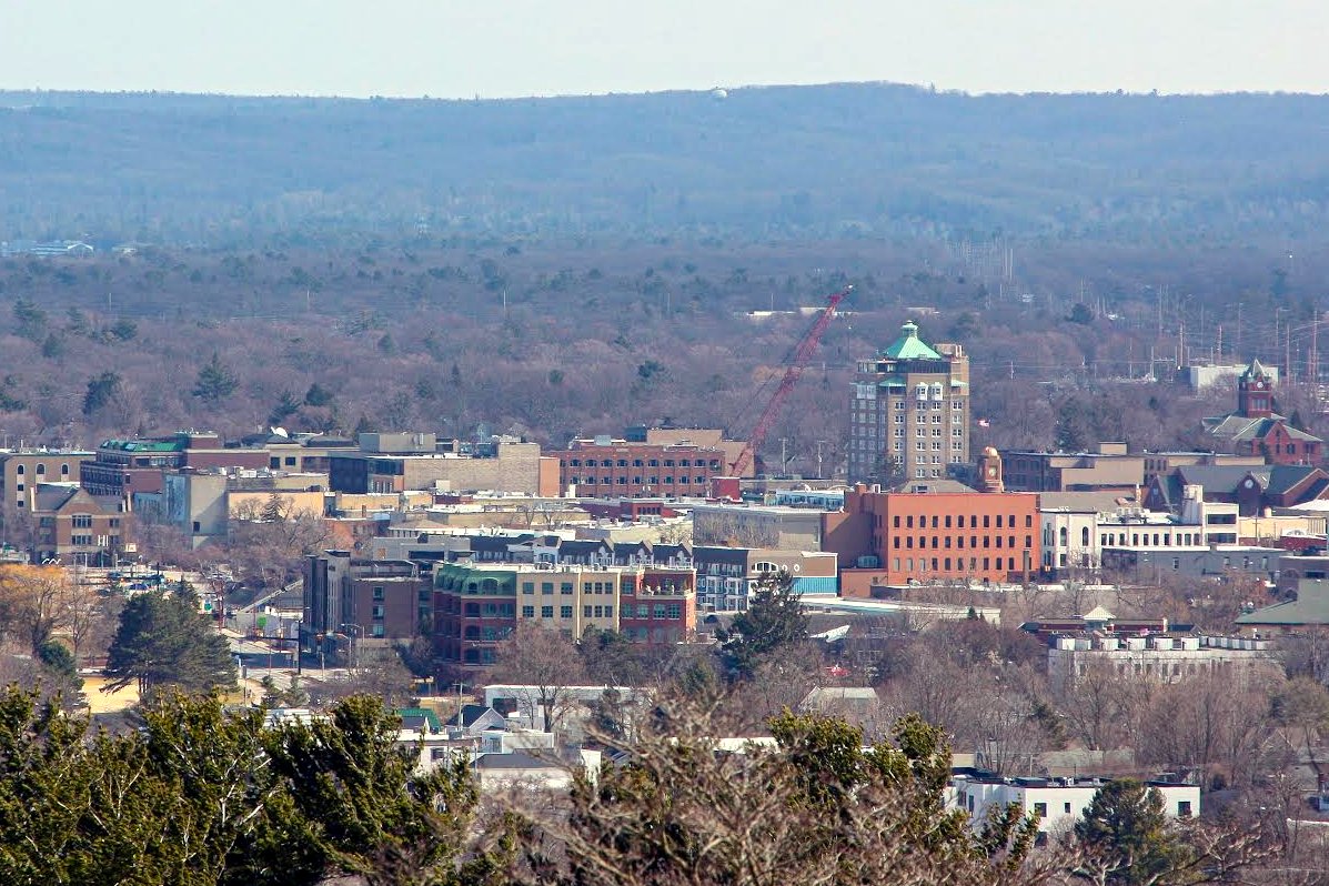 Downtown Traverse City’s low brick buildings and a tall white tower with a green cupola spread toward a wooded ridge under a hazy sky, seen from a hill, in a file photo.