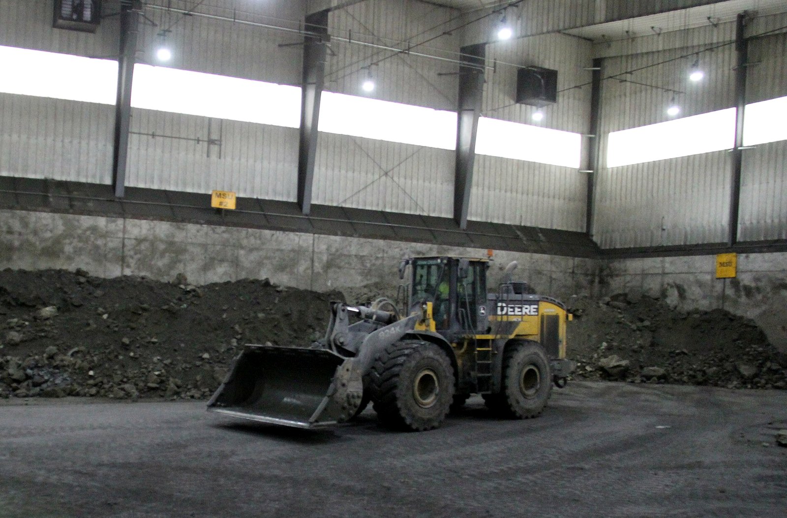 A yellow and black front-end loader with an empty bucket drives across a dark gravel floor inside a tall industrial building, with piles of broken rock against a concrete wall behind it.