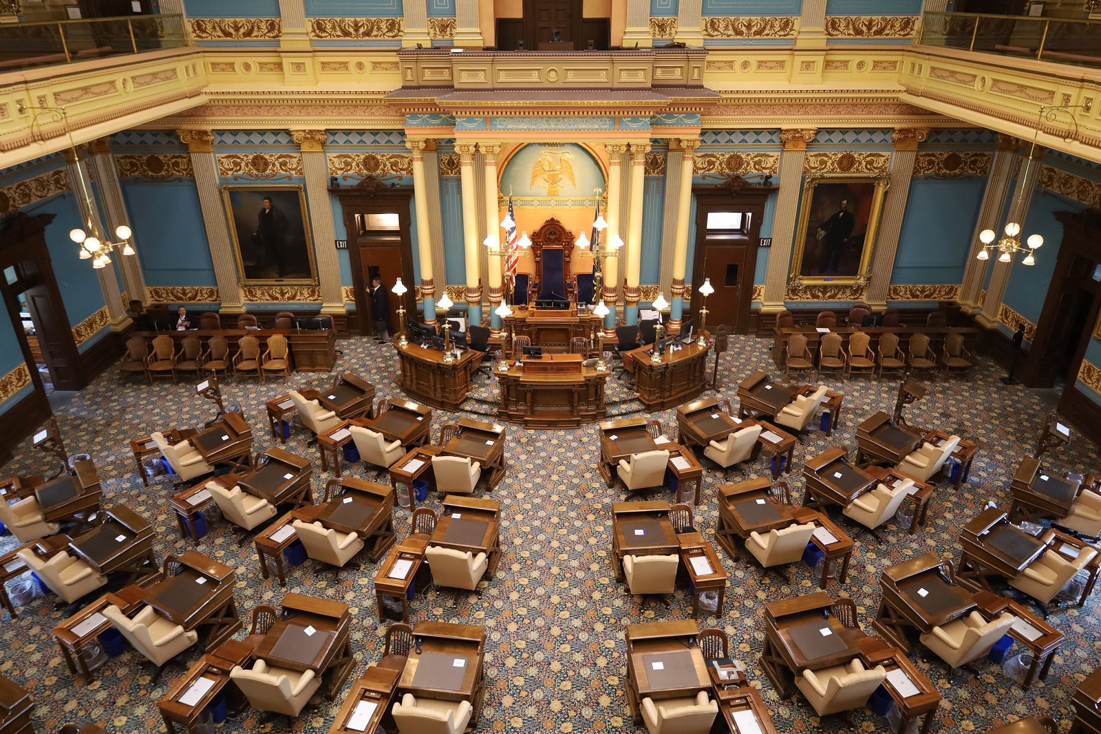 The Michigan Senate chamber in the State Capitol, seen from the gallery, with rows of wooden desks and tan chairs facing the rostrum under blue walls and gold trim.