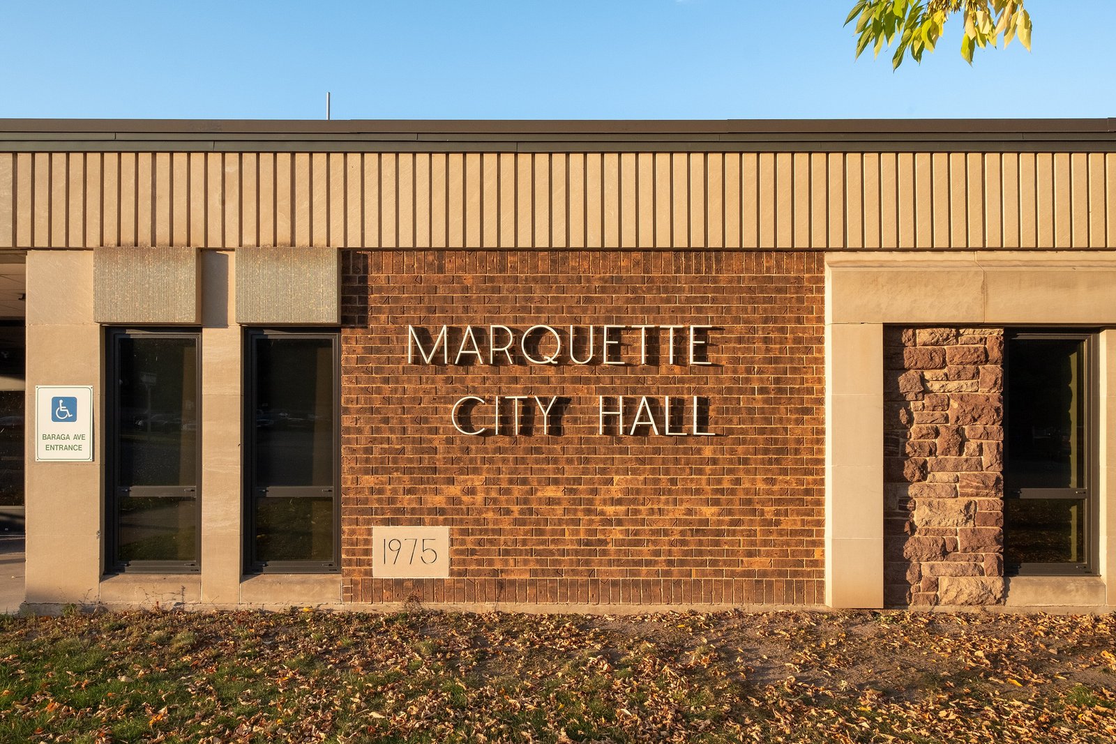 A tan brick wall with raised letters reading Marquette City Hall beside a row of dark windows, with lawn in front.