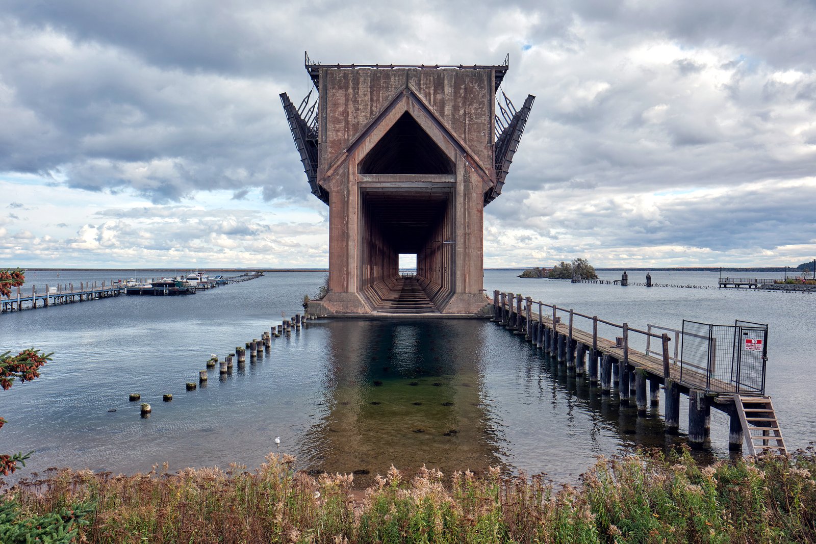 A tall concrete ore dock stands in calm harbor water under a cloudy sky, with a wooden pier on the right and tall grass in the foreground.