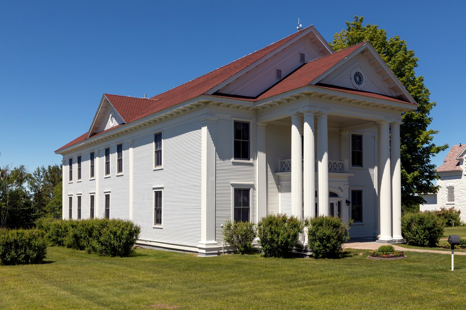 A white two-story courthouse with tall columns and a red roof sits on a green lawn under a deep blue sky, framed by shrubs and trees.