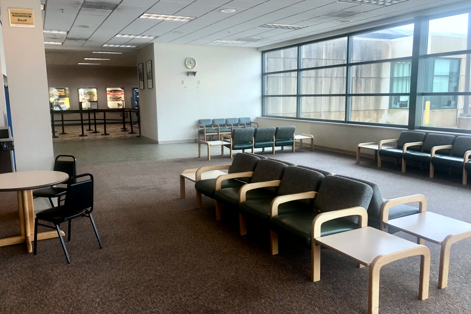 Rows of green upholstered chairs and small tables sit beside tall windows in a quiet airport terminal lounge.