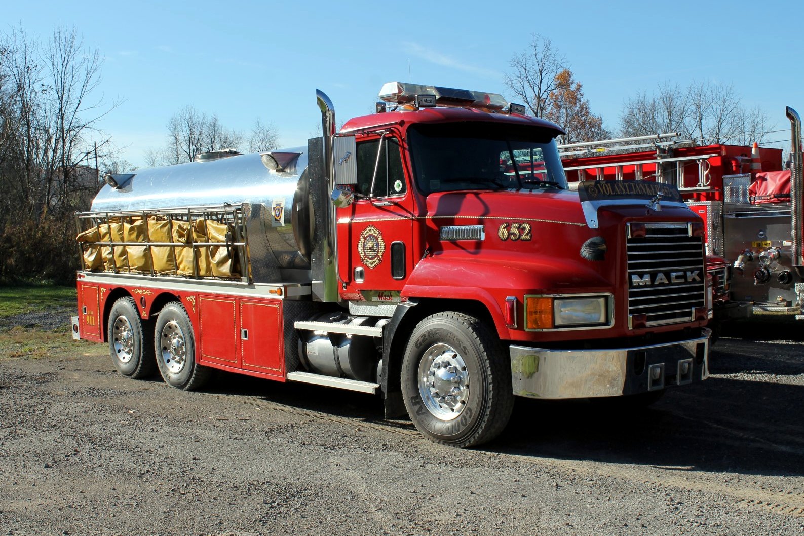 A red fire department tanker truck with a stainless-steel water tank and yellow hose bundles parked on gravel in front of other fire trucks.