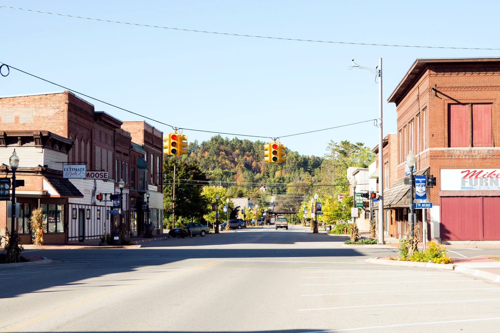 A wide, empty Main Street in downtown Norway, Michigan, on a sunny fall day, lined with two-story brick storefronts and corn-stalk decorations on the light poles, with traffic signals overhead and a hillside of green and orange trees rising at the end of the street.