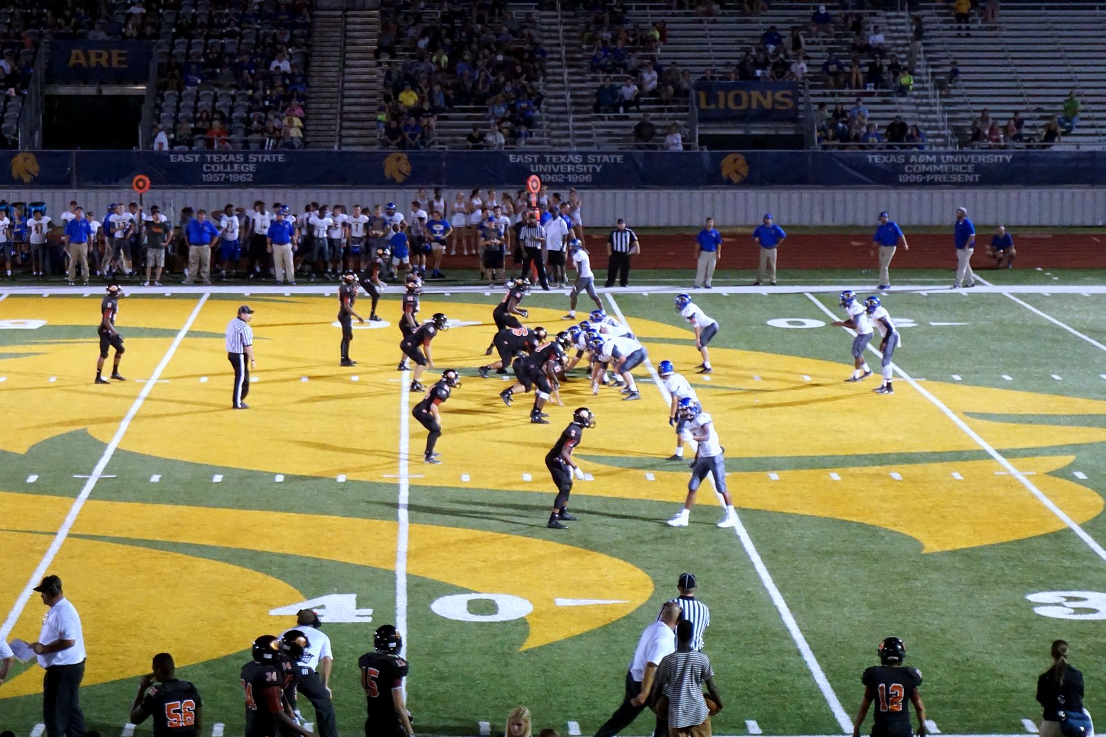 High school football players in dark and white uniforms line up at the line of scrimmage on a green-and-gold field at night as players and coaches watch from the sideline.