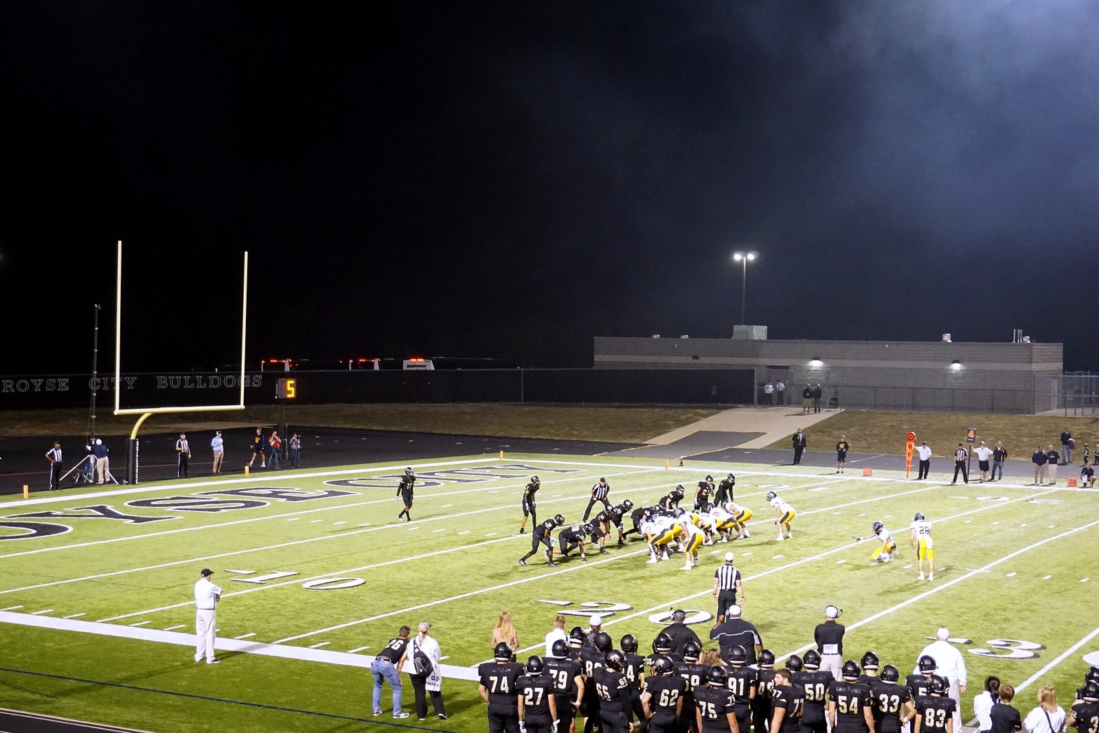A high school team lines up to kick a field goal at night on a lit turf field, with players in dark jerseys watching from the near sideline.