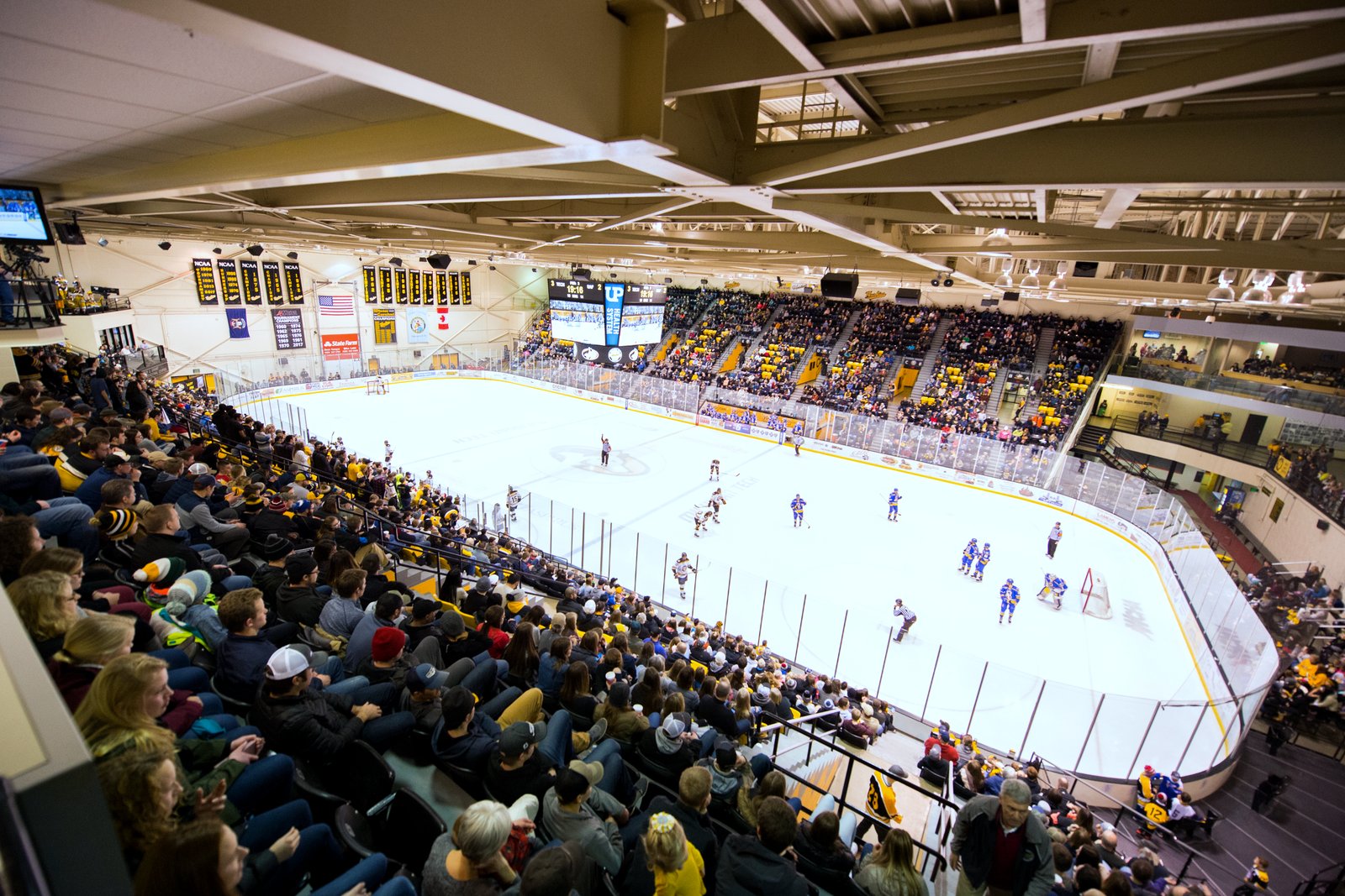 A wide view from the upper seats of a packed hockey arena, with fans in black and gold in the foreground and players in dark and blue jerseys on the white ice below.