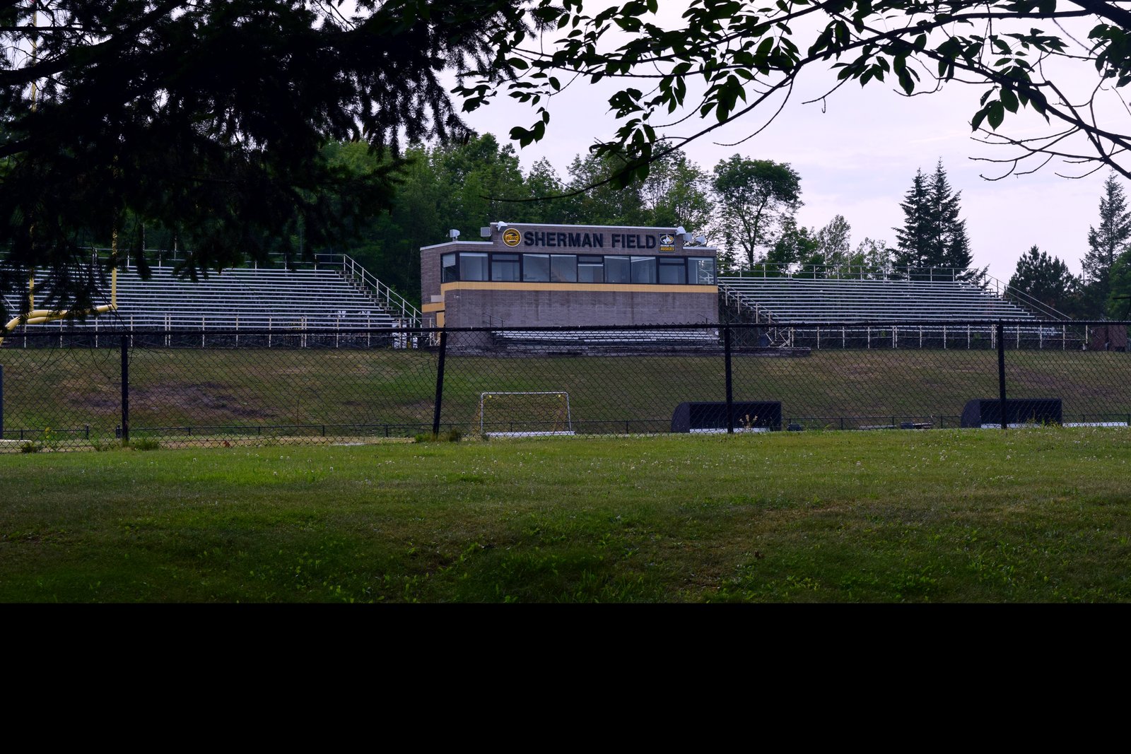 Sherman Field at Michigan Tech, with empty aluminum bleachers on both sides of a brick press box marked Sherman Field, seen through a chain-link fence and tree branches in summer.
