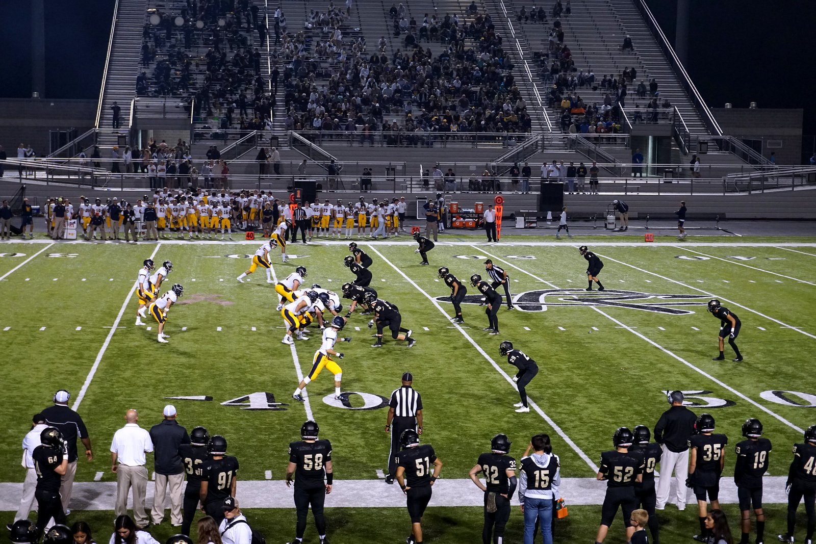 Two high school football teams line up for a play on artificial turf at night as players in black jerseys watch from the sideline and fans fill the bleachers.