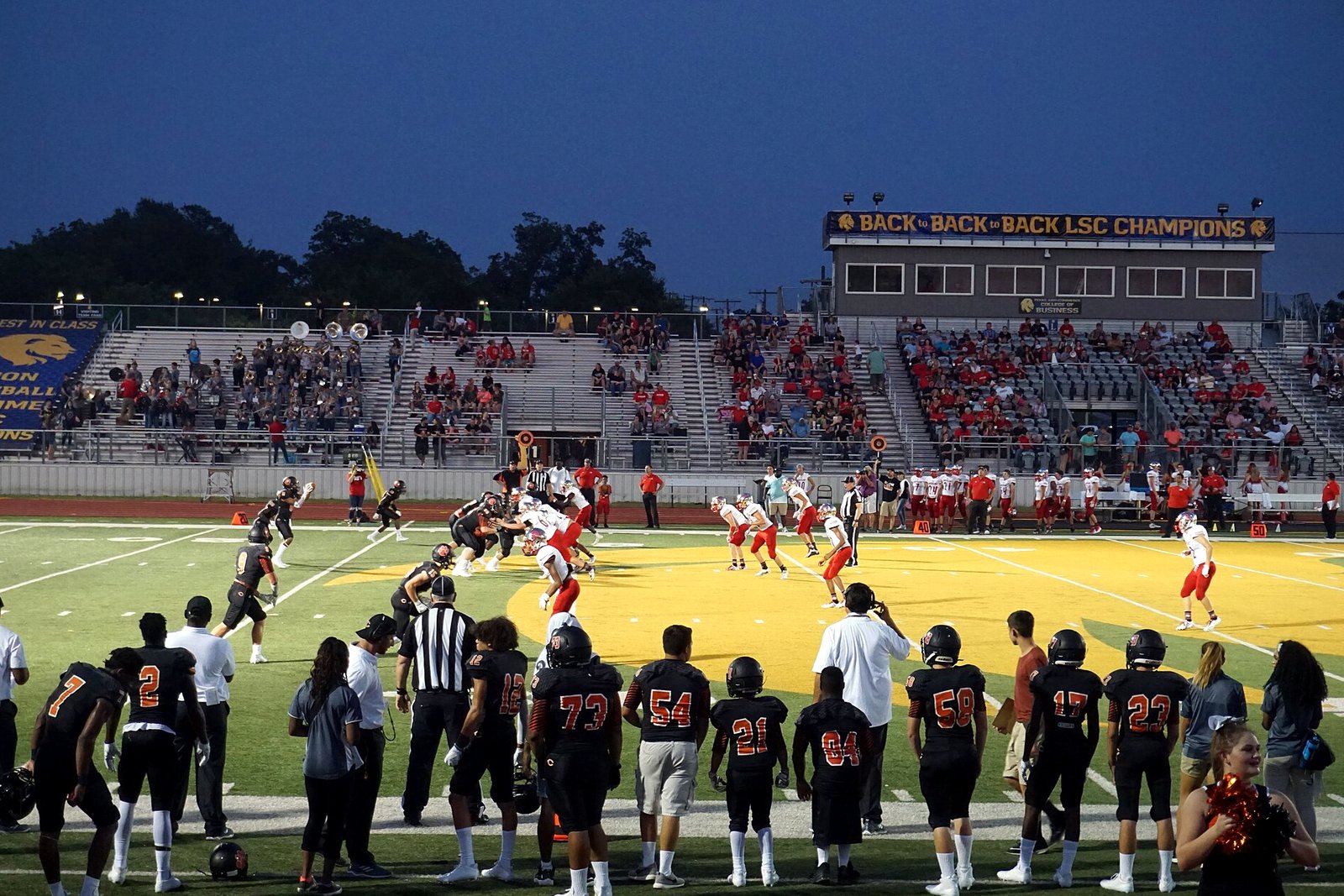 A high school football game on a gold field in front of crowded grandstands, with players in dark jerseys watching from the sideline.