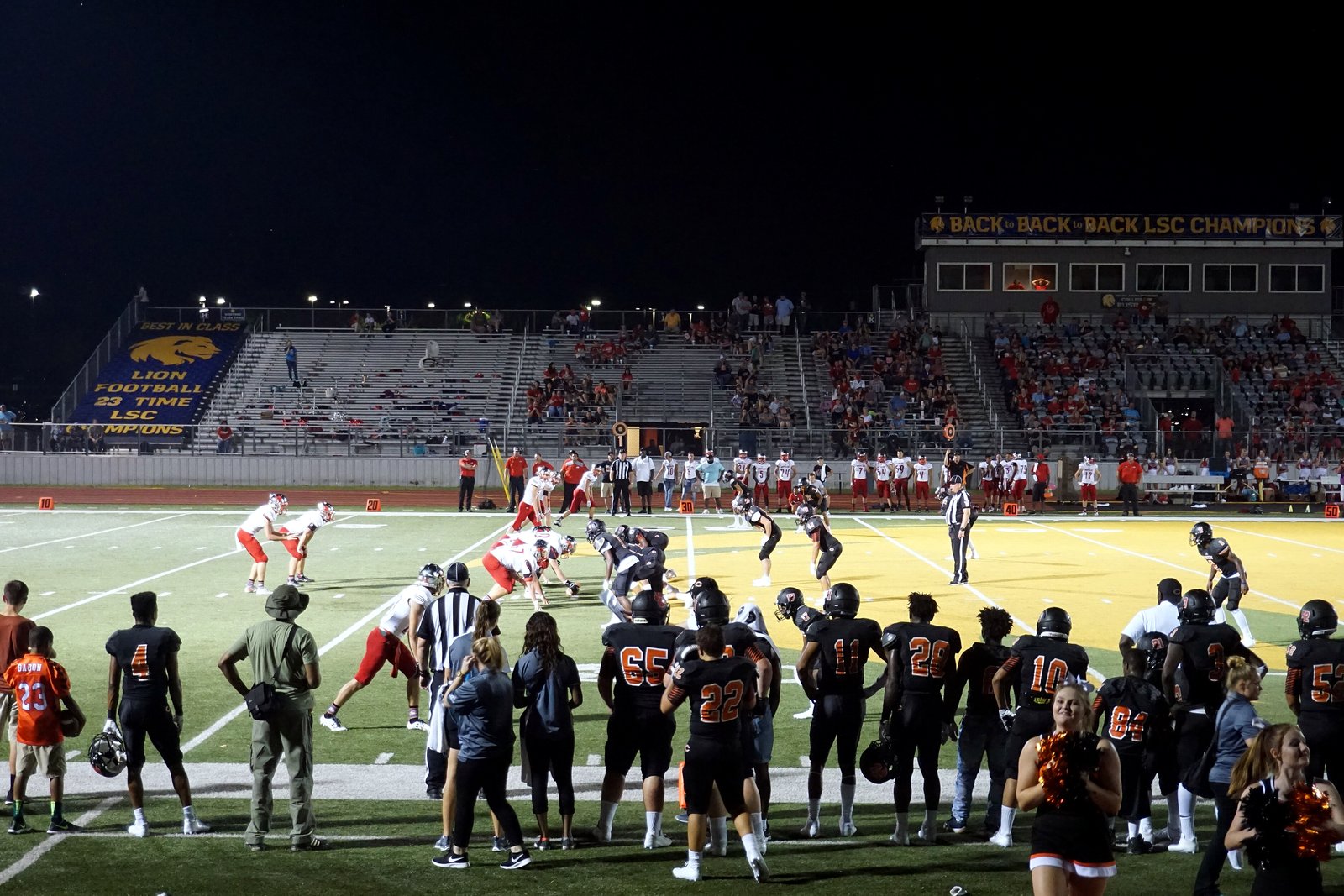 A high school football team in dark orange and black stands along the sideline at night as players run a play on the lit field in front of the home stands.