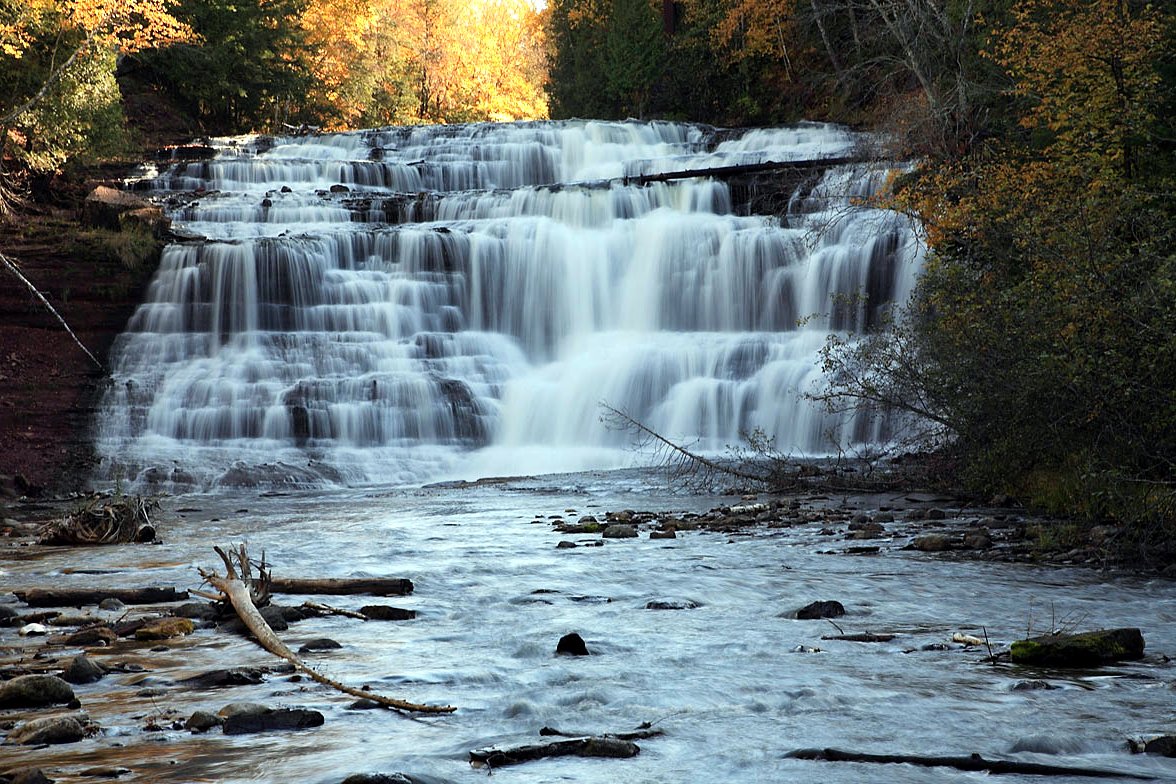 A wide, terraced waterfall spills over layered rock into a shallow, rocky river, framed by orange and green fall trees.