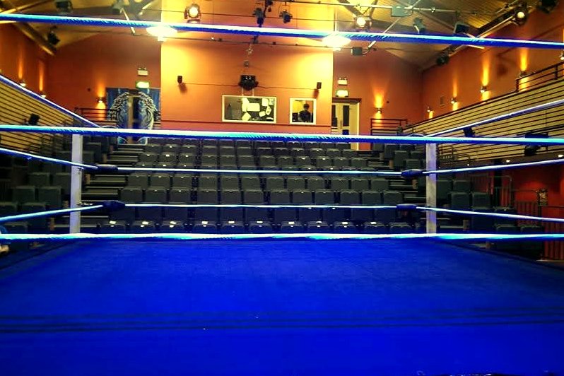 A professional wrestling ring with a blue mat and ropes set up on the floor of an empty theater hall with tiered seats.