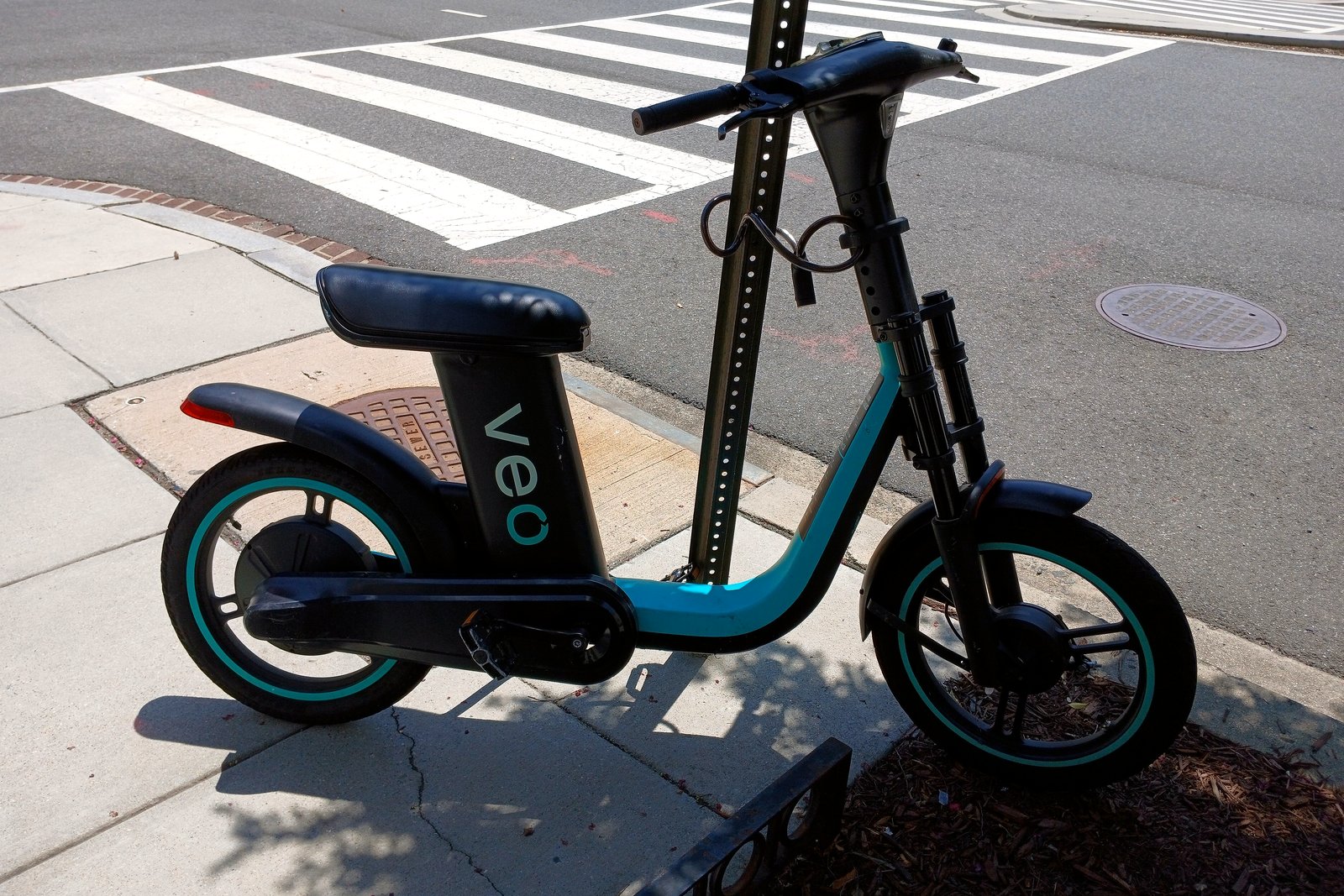 A parked teal and black electric bike leaning beside a signpost on a city sidewalk near a crosswalk.