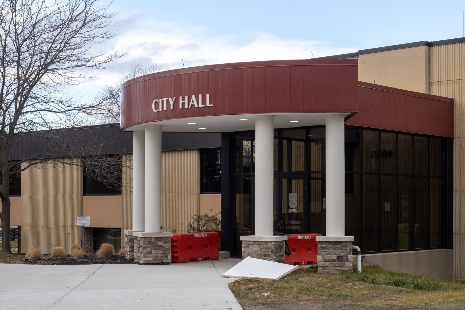A one-story tan and dark brown municipal building with a curved maroon canopy reading CITY HALL over a columned entrance, with bare trees and a walkway in front.