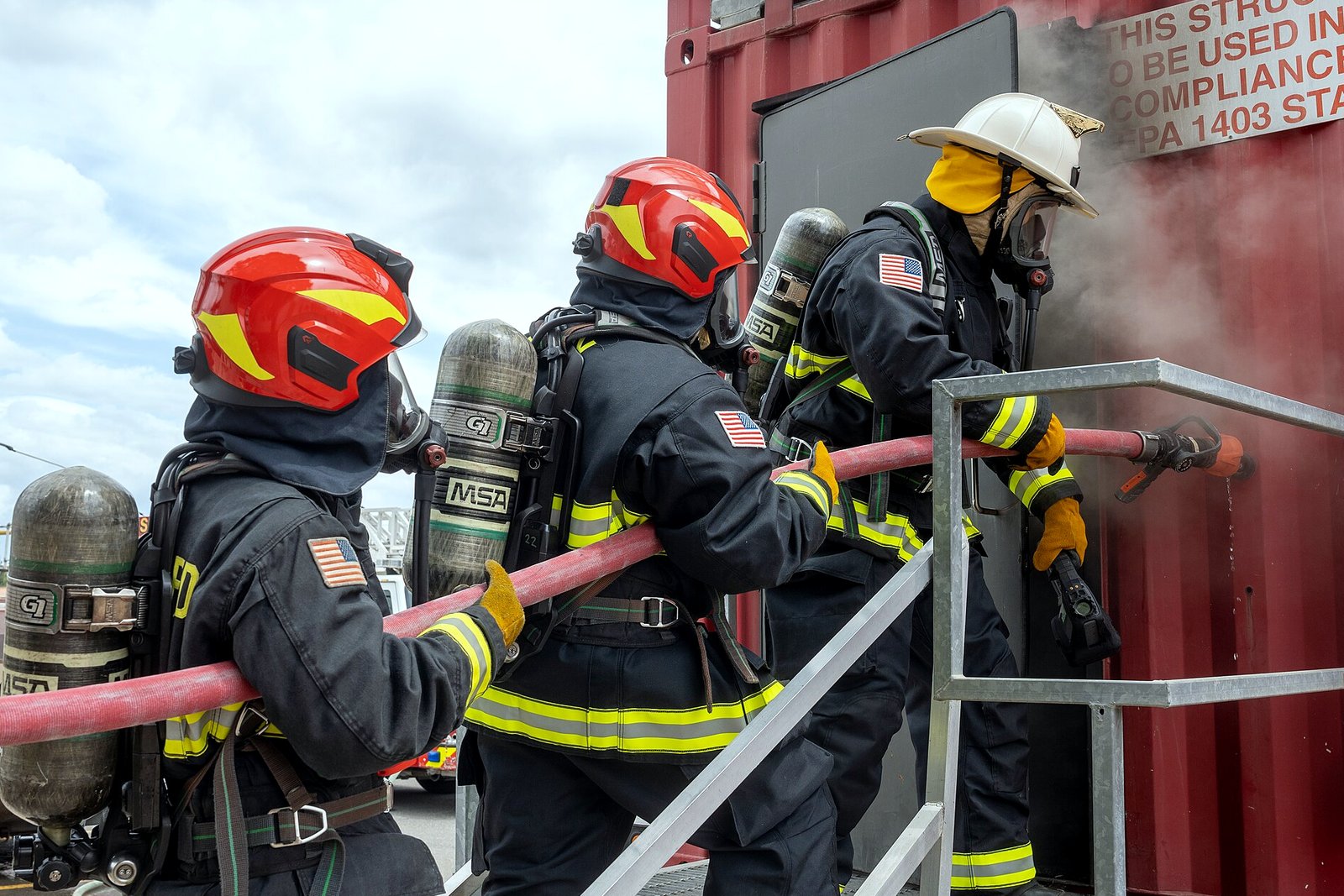 Three firefighters in full protective gear and air tanks move a charged hose line into a smoking red steel training container.