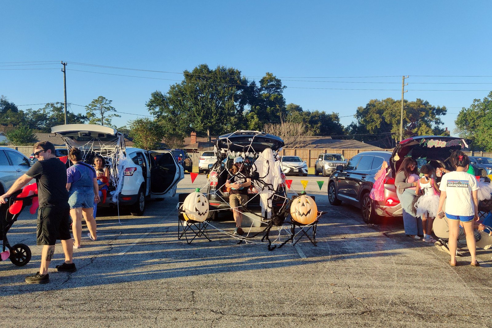 Families in costumes walk among decorated cars with open trunks in a church parking lot on a sunny fall afternoon.