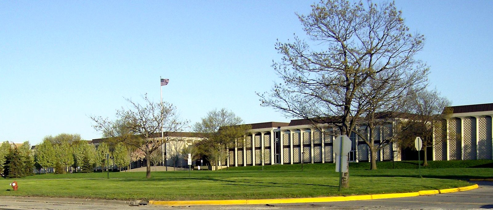 A long, low brick and concrete college building behind a wide lawn with bare trees and a flagpole under a blue sky.