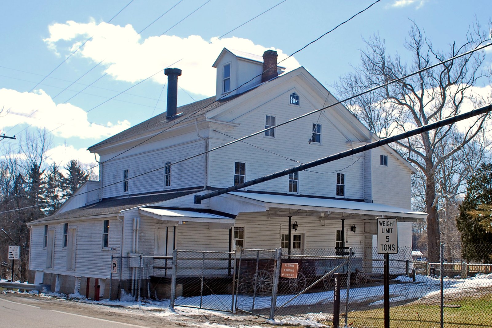 A white, two-and-a-half-story wooden mill building with a porch and a metal roof beside a road, with patches of snow on the ground and bare trees behind it.