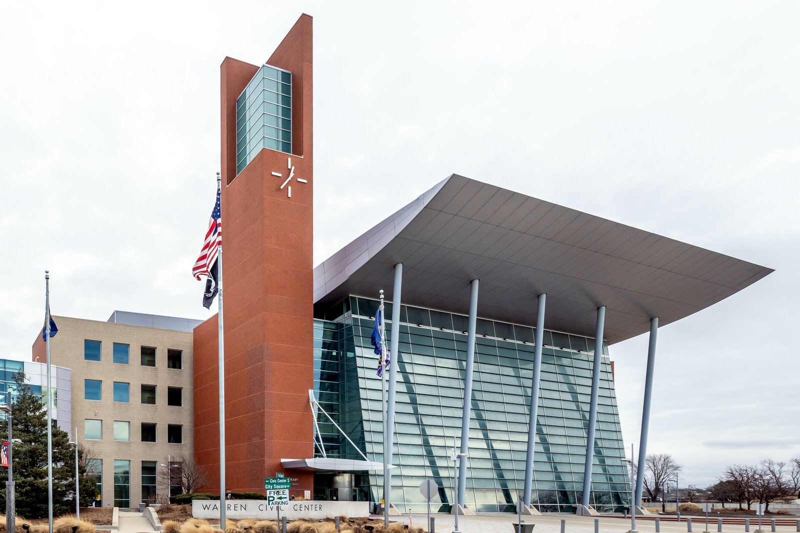 A tall red-brick clock tower beside a glass building under a wide, sweeping metal canopy, with flags out front and a Warren Civic Center sign along the walk.