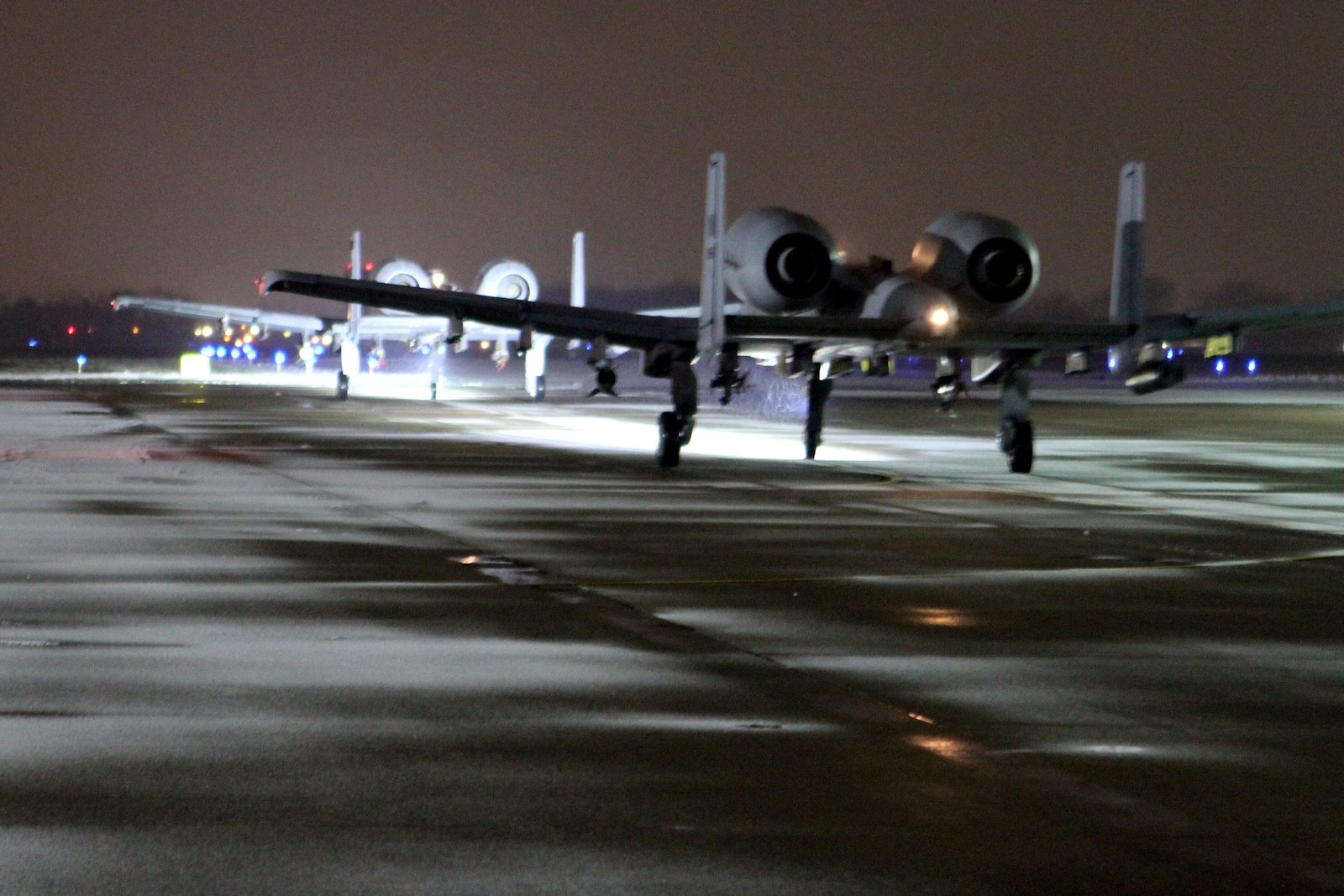 Two gray A-10 attack jets taxiing toward the viewer on a wet, lighted airfield at night.