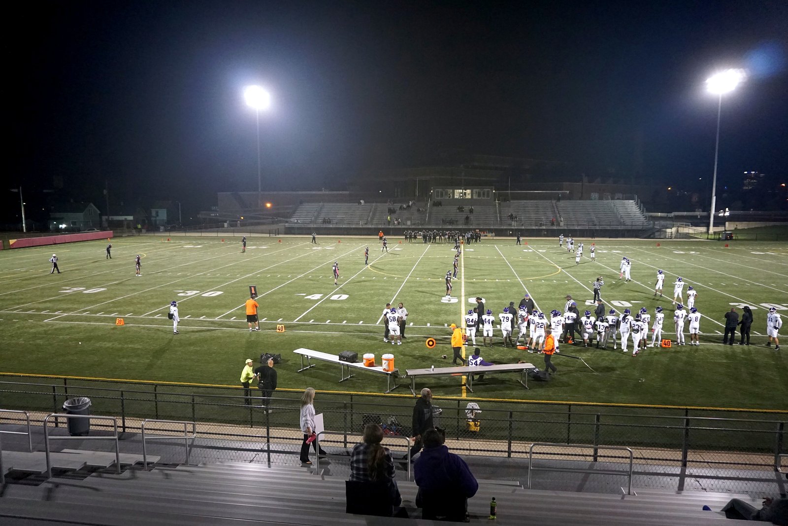 A high school football team kicks off at night on a lit turf field, seen from the stands behind a railing, with light towers and bleachers across the field.