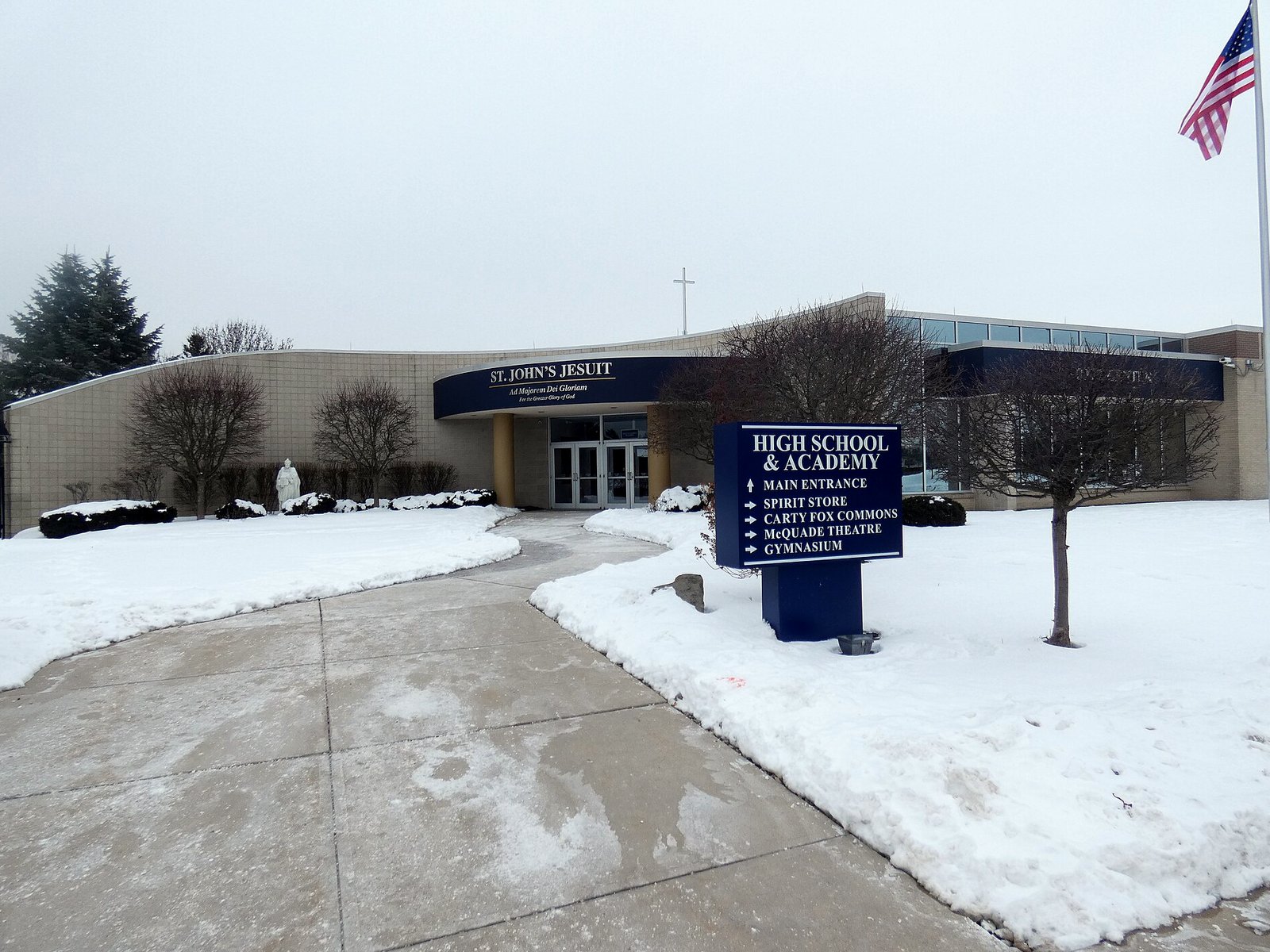 The curved, cream-colored front of a school with a navy awning reading St. John's Jesuit, a blue sign listing the high school and academy entrances, snow-covered lawns, bare trees and an American flag on a pole, under a gray sky.