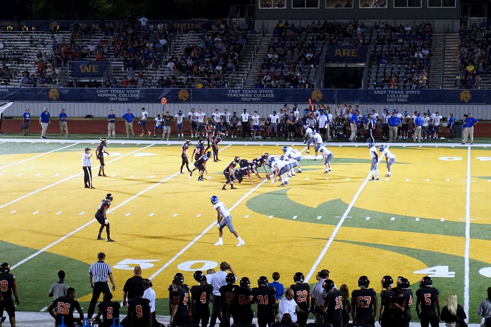 High school football players in white jerseys run an offensive play as defenders close in on a sunny field, in a file photo.