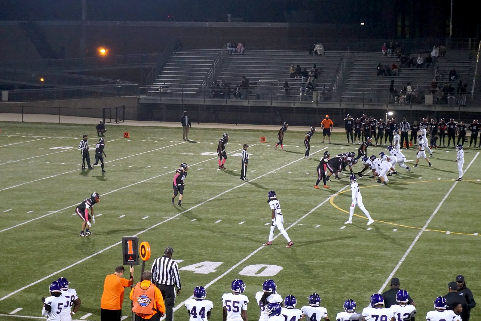 Under the lights, a team in dark jerseys and a team in white jerseys line up at the line of scrimmage on a turf field, with players in white jerseys and purple helmets on the sideline in the foreground.