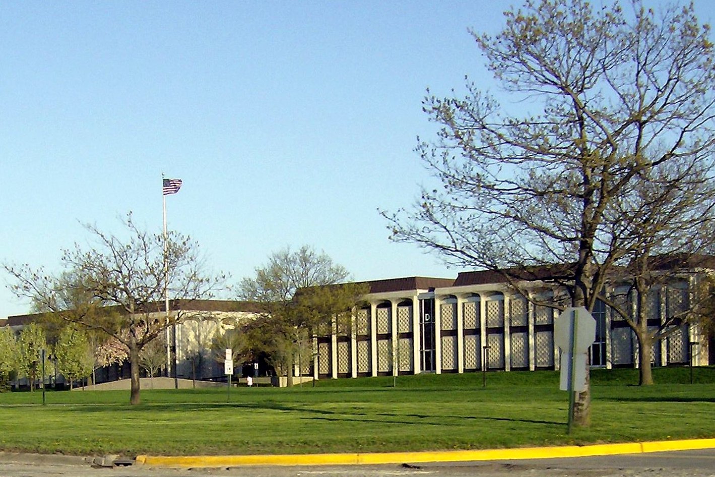 Long, low brick-and-concrete college buildings with patterned window columns sit behind a wide lawn, a flagpole and budding trees under a clear blue sky.