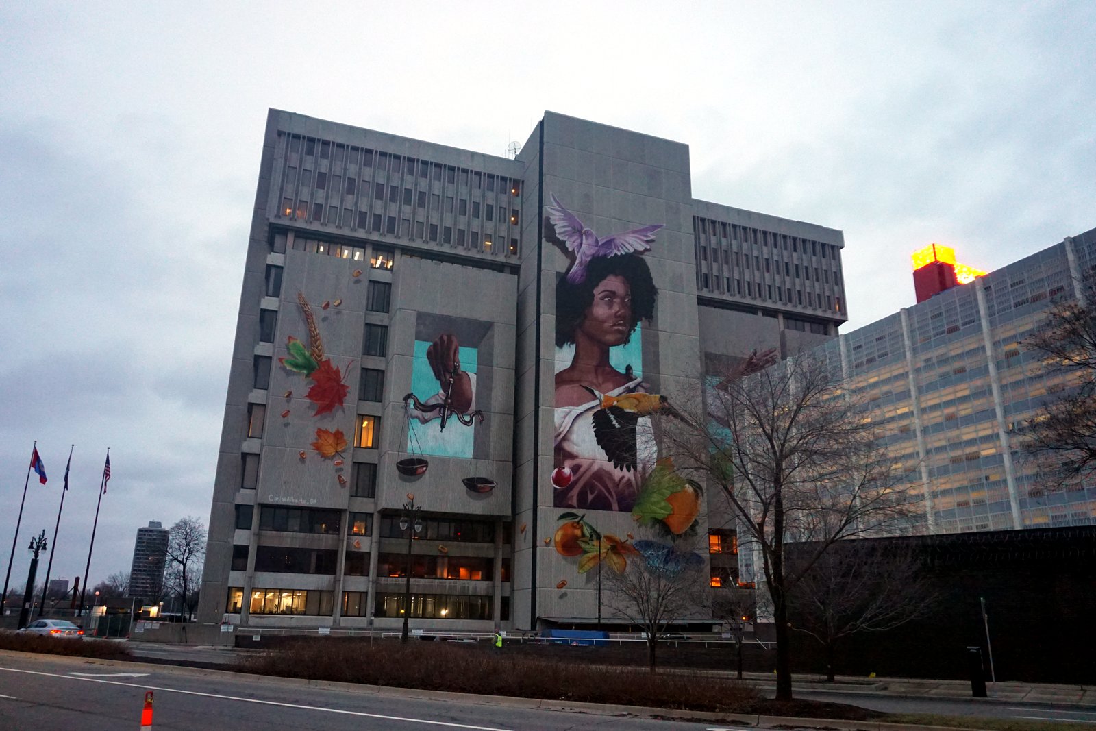 A tall, boxy concrete courthouse with a large colorful mural of a woman’s face on its side stands behind bare trees and flagpoles under a gray winter sky.